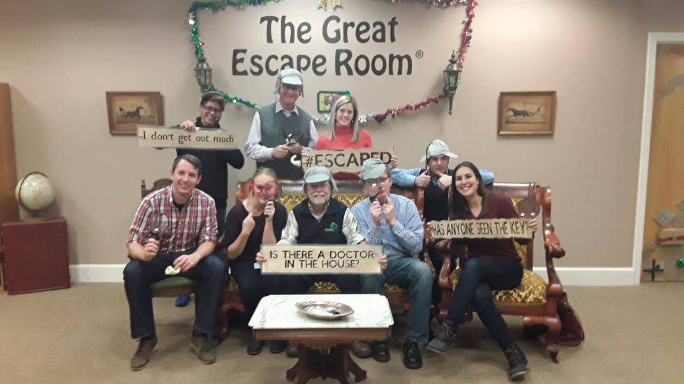 Photograph of a group of eight people posing in a room labeled "The Great Escape Room," holding humorous signs related to escape room themes. The setting includes vintage furniture, festive garlands, and signs with phrases like "I don't get out much," "ESCAPED," and "Has anyone seen the key?" highlighting a successful escape experience.