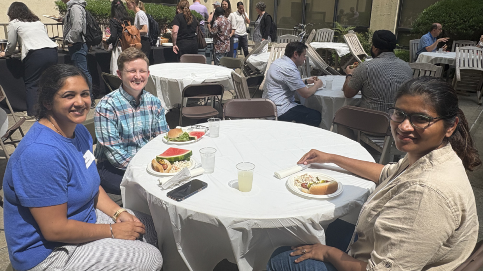 People seated at round tables outdoors in a courtyard beside tall building walls, eating and talking; several guests smile while others converse, with white tablecloths, plates, and drinks on the tables.