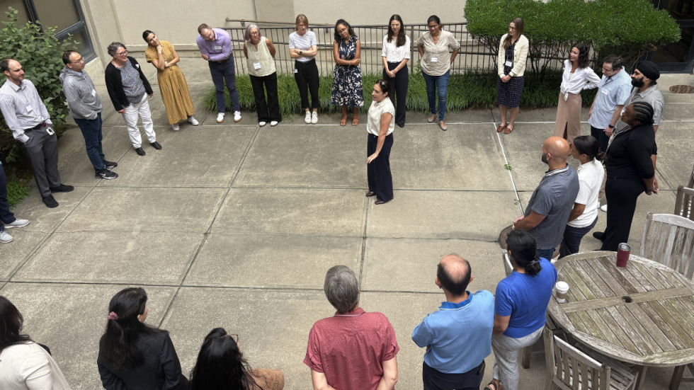 A group of people stand in a circle outdoors on a campus walkway near a building, with one person in the center speaking while others listen, photographed from above.