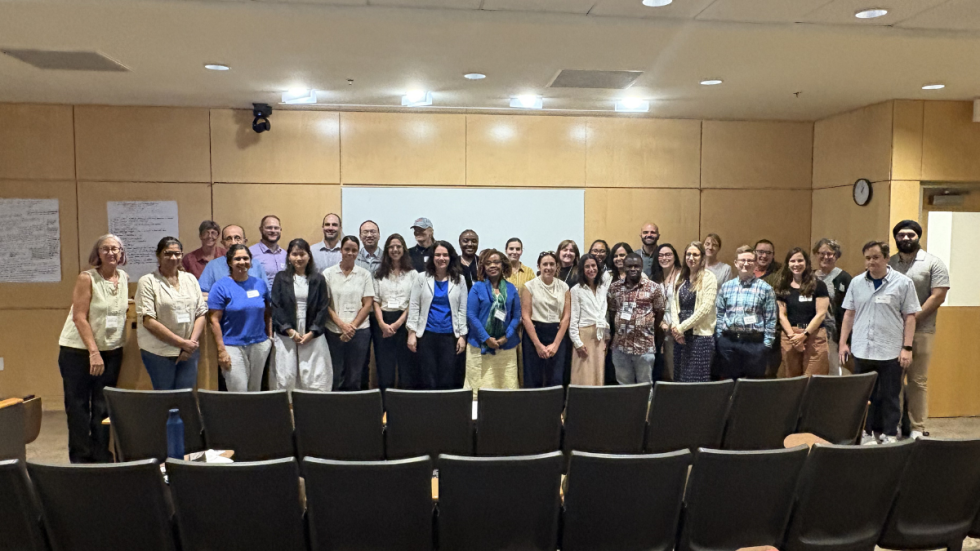 Group photo of about 31 people standing in a university-style lecture room in front of a projector screen, with empty black chairs in the foreground and a few papers and notebooks on seats.